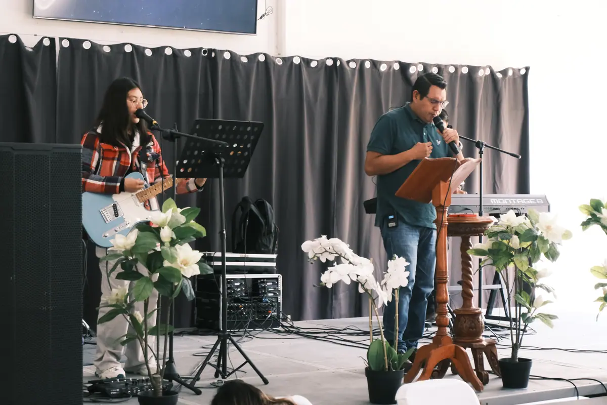 Una mujer canta con guitarra y un hombre habla desde el atril, con flores blancas al frente, durante un servicio.
