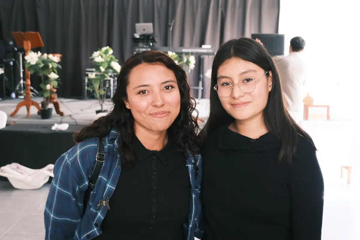 Dos jóvenes de la iglesia sonríen a la cámara, una junto a la otra, frente al escenario.