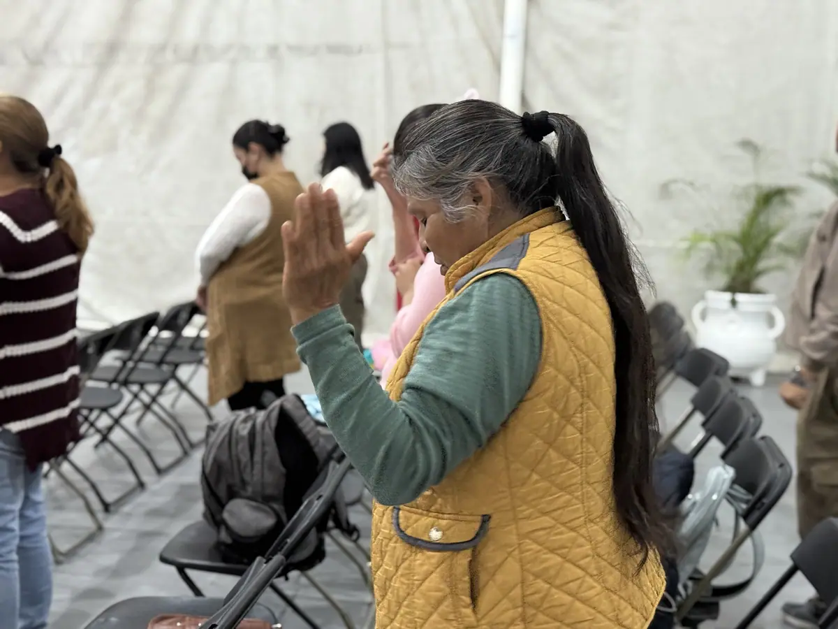 Una mujer mayor con chaleco amarillo ora con las manos juntas frente al rostro y los ojos cerrados.