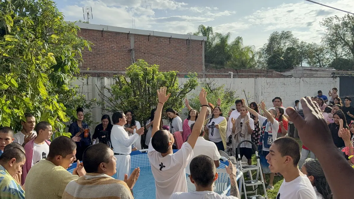 Personas de la congregación levantan las manos en adoración al aire libre, junto a una alberca, bajo los árboles.