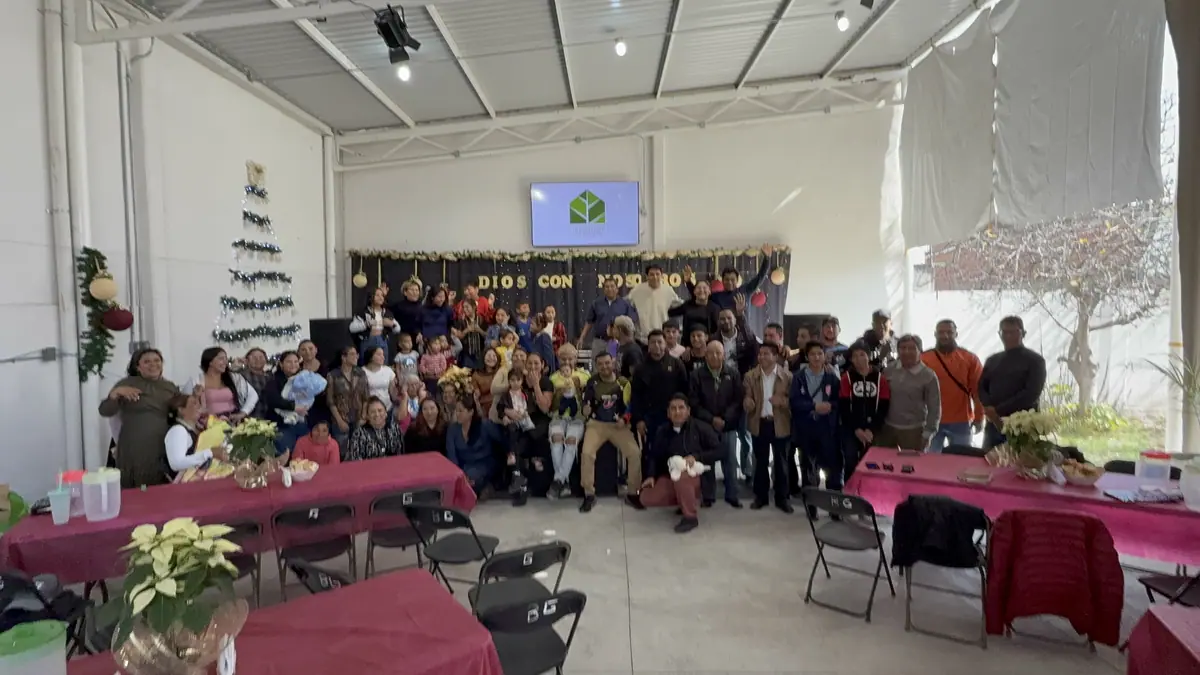 La congregación posa para una foto de grupo en el salón, bajo un letrero que dice «Dios con nosotros».