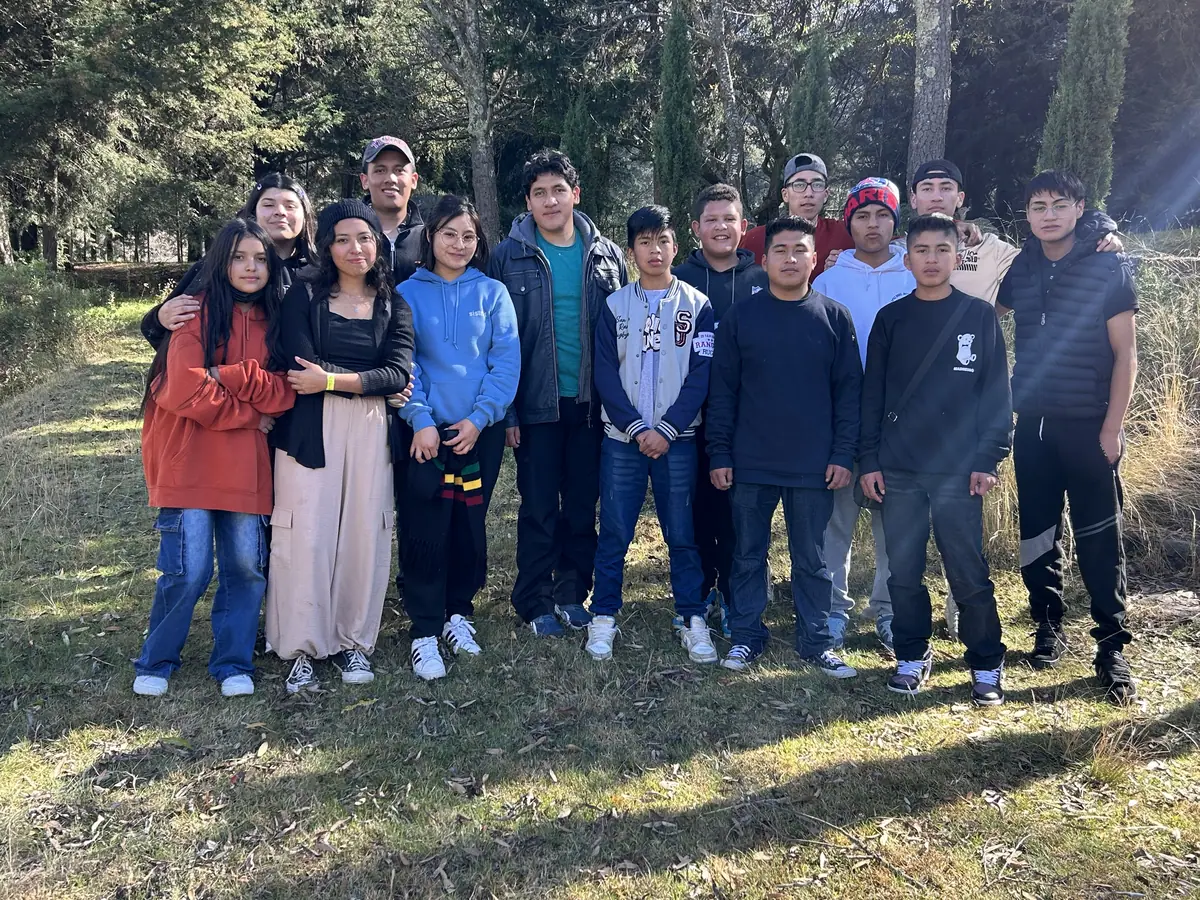 Un grupo de jóvenes y adultos posa junto, al aire libre, sobre el pasto y entre árboles.