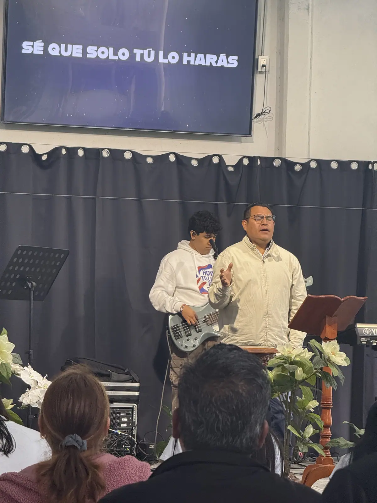 Un hombre habla desde el atril mientras un joven toca el bajo, con flores blancas al lado.