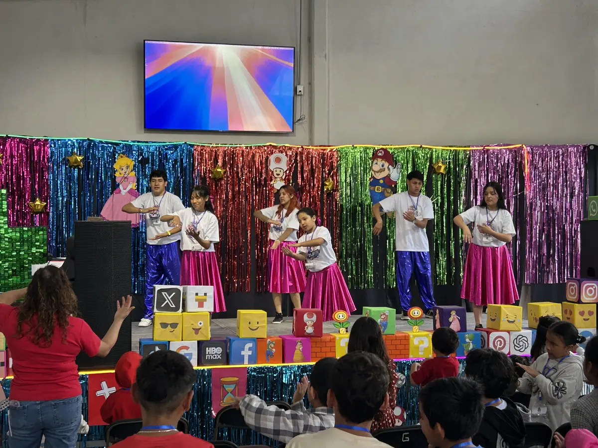 Niñas y niños con ropa de colores se presentan sobre un escenario decorado con cortinas brillantes.