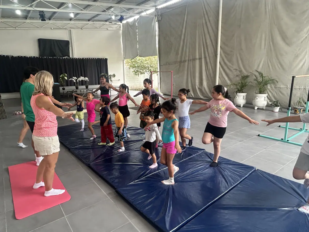 Niños y niñas recorren un circuito de ejercicios sobre colchonetas, guiados por una mujer.