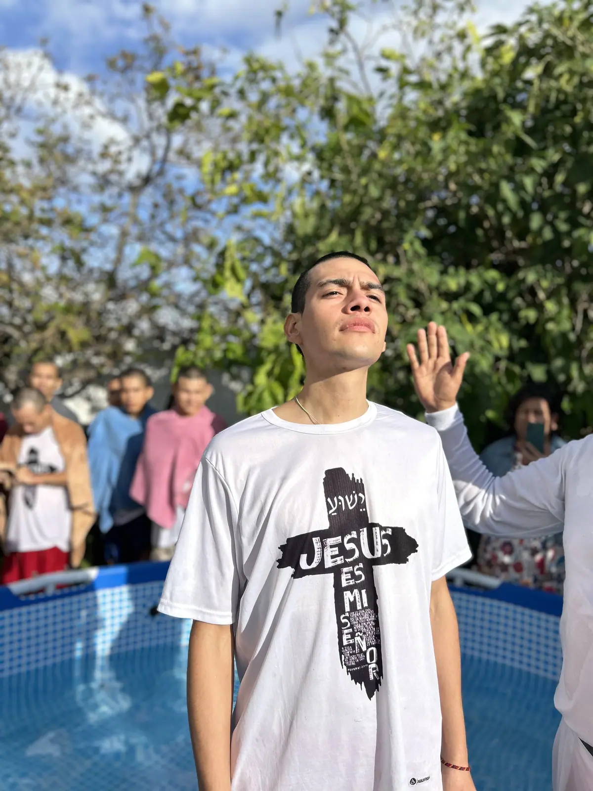 Un joven con una camiseta blanca con una cruz y las palabras Jesús es mi Señor, dentro de una alberca durante su bautismo, mientras un hombre vestido de blanco levanta la mano sobre él.